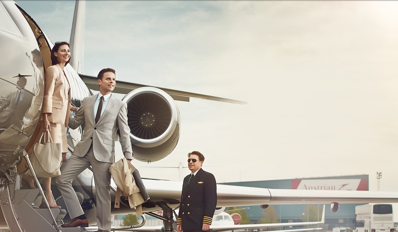 : Elegantes Paar steigt aus einem Privatjet, Flughafenmitarbeiter steht im Hintergrund.